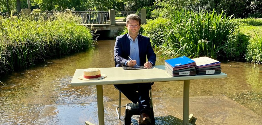 Bürgermeister Christoph Glogger hat seinen Arbeitsplatz in die Isenach Bad Dürkheim verlegt und sitzt am Schreibtisch im Gewässer.