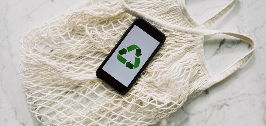 Symbol für nachhaltige Beschaffung: Einkaufstasche aus Garn mit Handy im Mittelpunkt, auf dem das Symbol für Nachhaltigkeit zu sehen ist