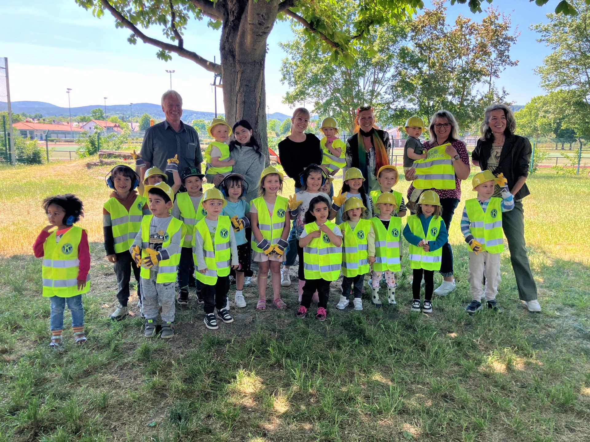 Eine Gruppe von Kita-Kindern steht mit Erwachsenen unter einem Baum auf einer Wiese. Die Kinder tragen gelbe Bauhelme, Warnwesten und Arbeitshandschuhe. Sie lächeln in die Kamera. Im Hintergrund sieht man Bäume, einen Zaun und Gebäude.