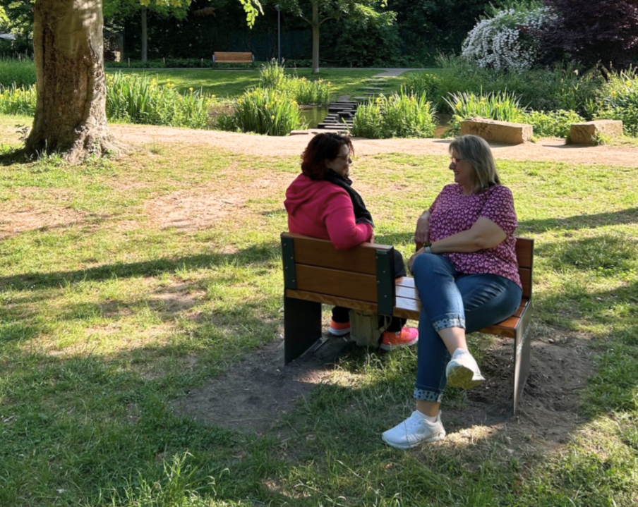 Im Kurpark sitzen zwei Frauen auf einer doppelseitigen Holzbank einander zugewandt und unterhalten sich lächelnd. 