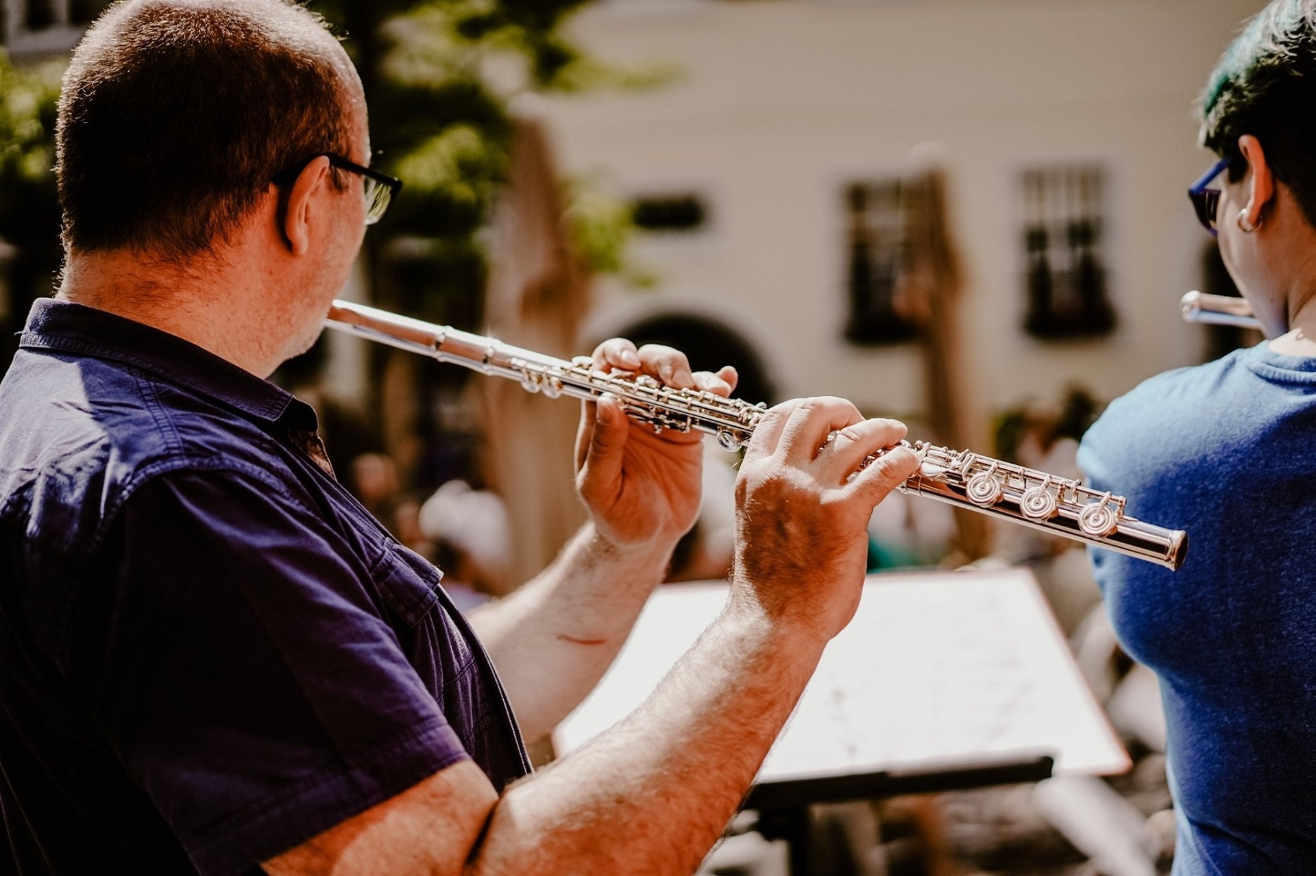 Querflötenspieler Zu sehen ist ein Mann von hinten, der Querflöte spielt. Er blickt von der Kamera weg auf einen Notenständer. Unscharf ist eine weitere Person zu sehen