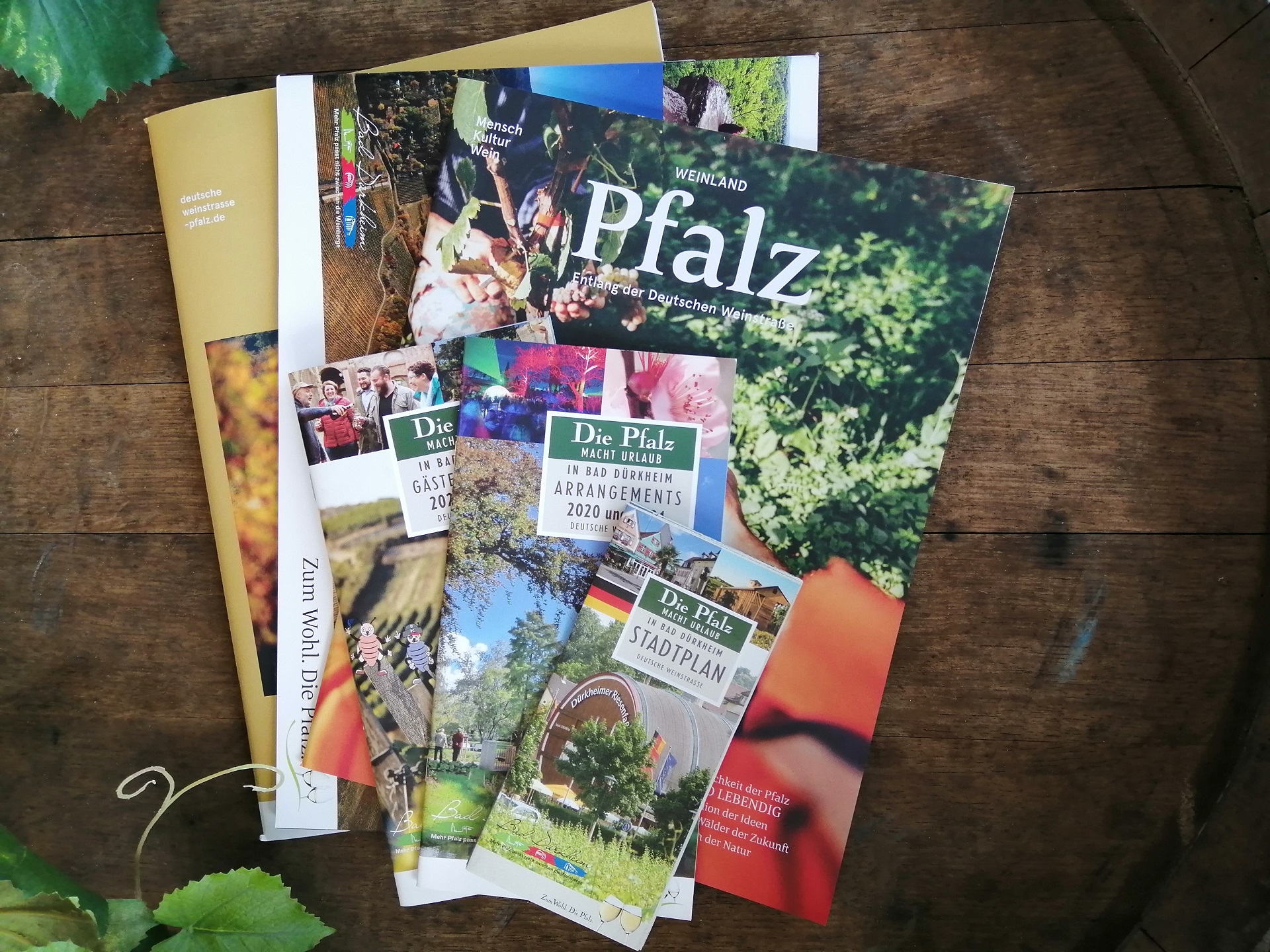 Informationspaket Bad Dürkheim Prospekte von Bad Dürkheim die im Informationspaket enthalten sind, auf einem Faß dargestellt, umrahmt werden die sechs Produkte mit Weinreben in der linken oberen und unteren Ecke des Bildes.