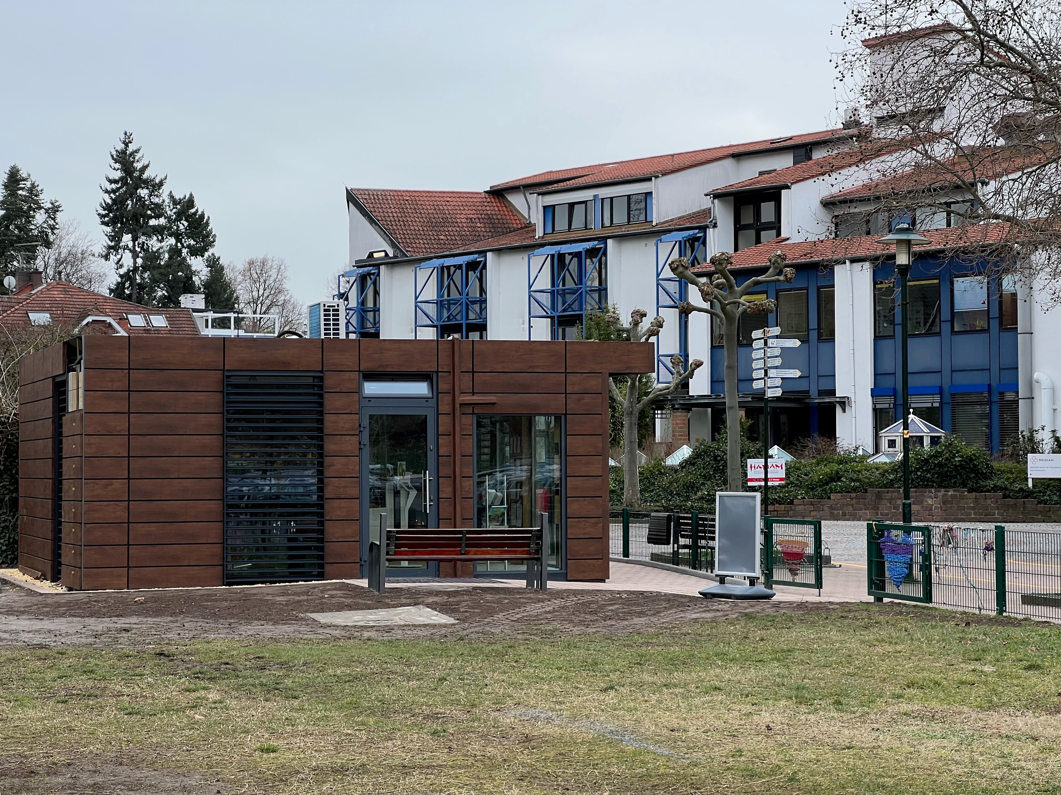 Ein Container in dunkelbrauner Holzoptik, im Vordergrund ist ein kurzer Teil einer Wiese zu sehen. Im Hintergrund die Median Klinik für Psychosymatik.