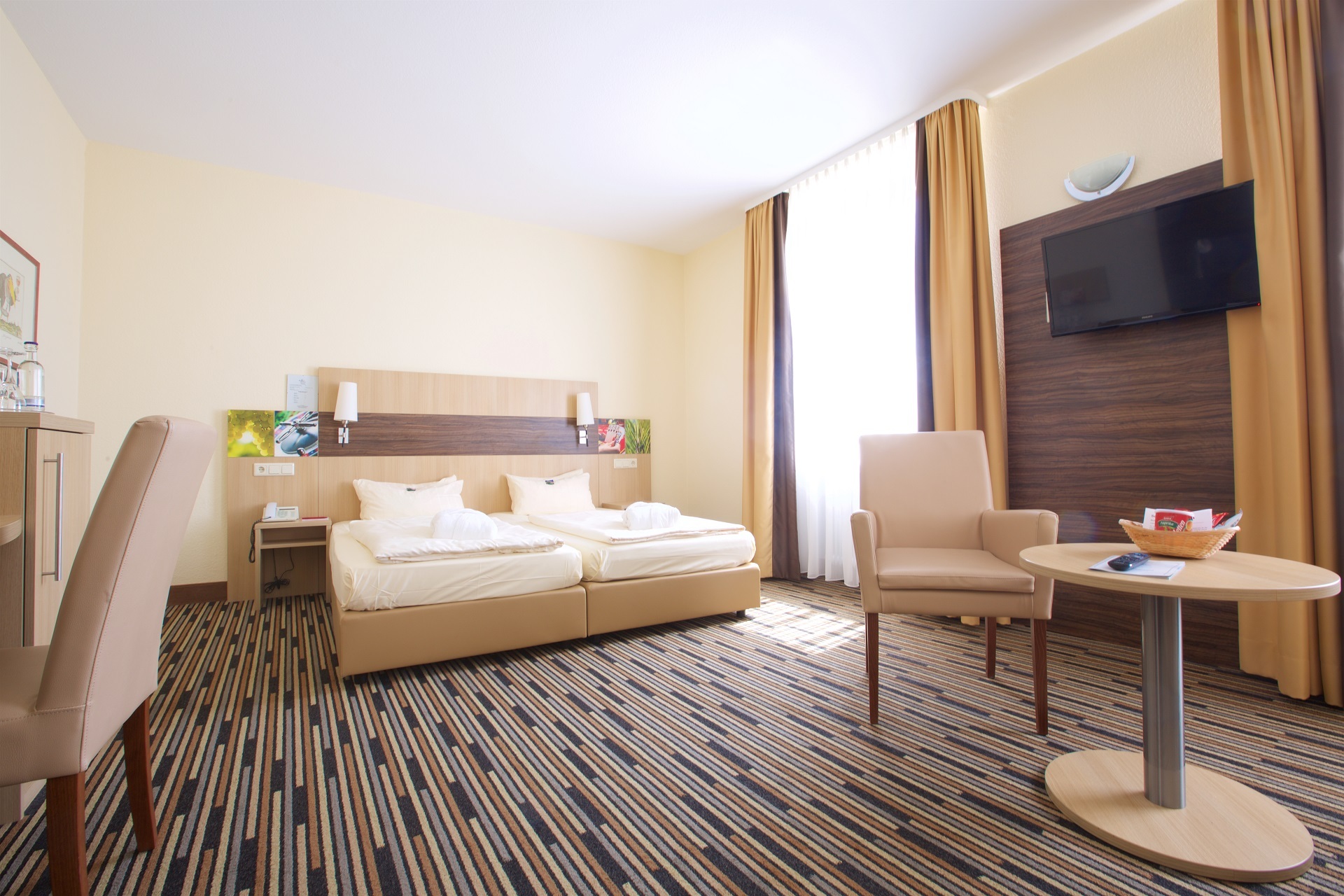 Auf dem modernen gemusterten Teppich steht eine helle Hotelzimmerausstattung mit Schreibtisch Bett und Fernseher. Im Hintergrund auf der rechten Seite ein Fenster.