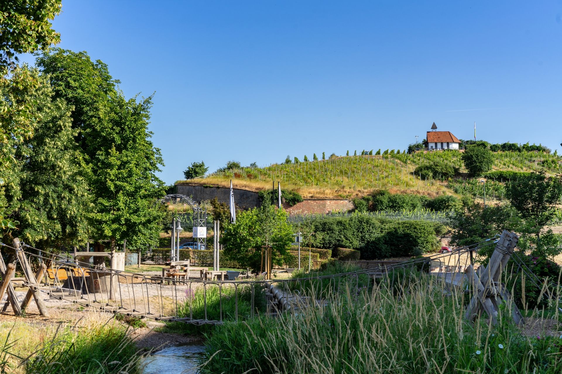 Durch das Schilf im Vordergrund an der Isenach, blickt man auf die Spielgeräte des Wasserspielplatzes die mit Sand umgeben sind. Überhalb im Hintergrund thront die Michaeliskapelle auf dem Weinberg.