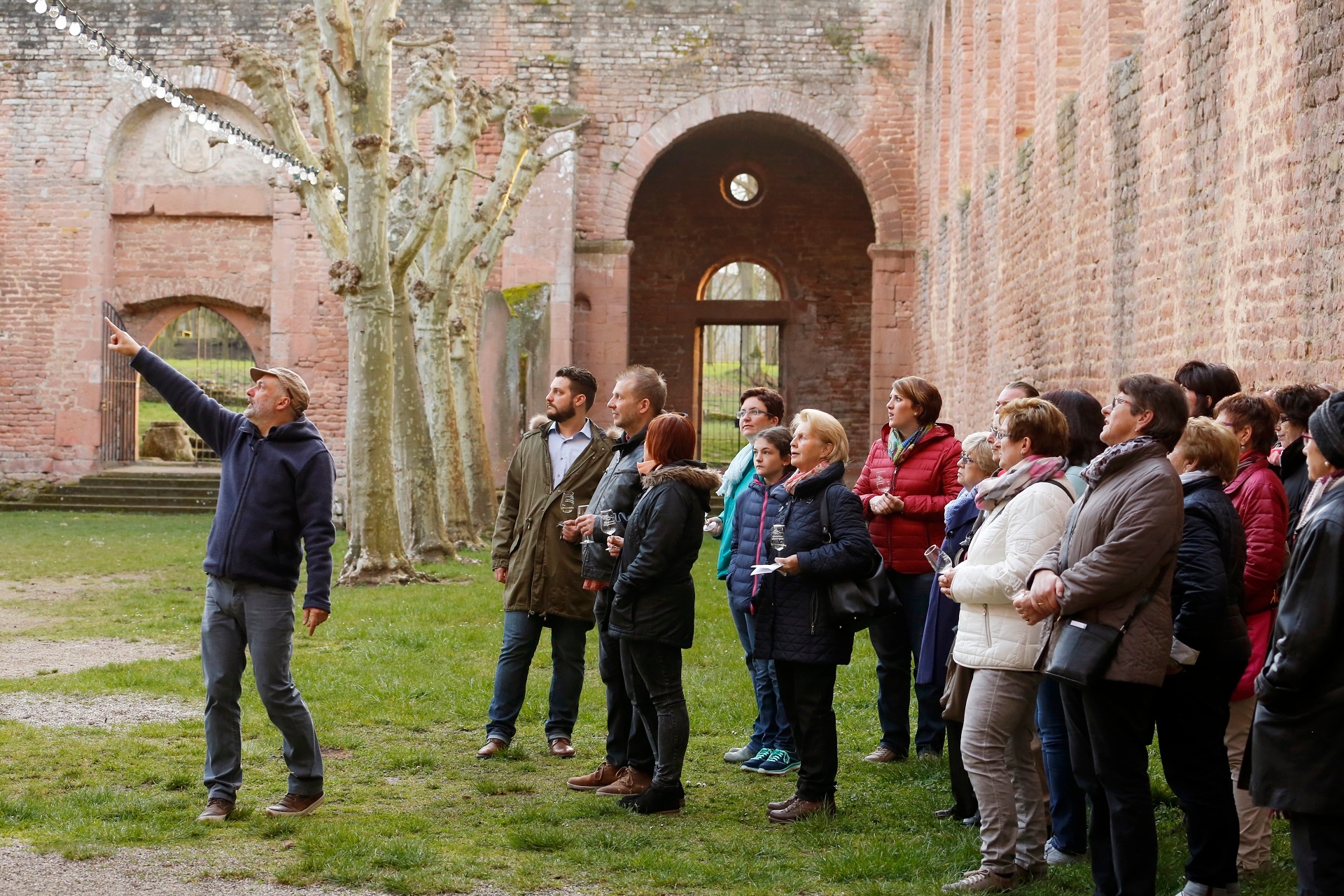 Führung in der Klosterruine Limburg Rechts im Bild eine Gruppe die dem Gästeführer links aufmerksam zuhört, dieser deutet gerade mit seinem ausgestreckten Finger auf die Ruine. Die Gruppe befindet sich in der Klosterruine Limburg, sie ist im Hintergrund zu sehen.