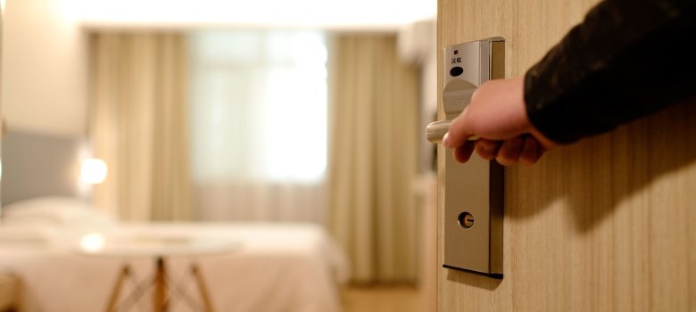 Eine Hand öffnet eine Türe in ein Hotelzimmer, der Blick fällt auf das Bett im Zimmer
