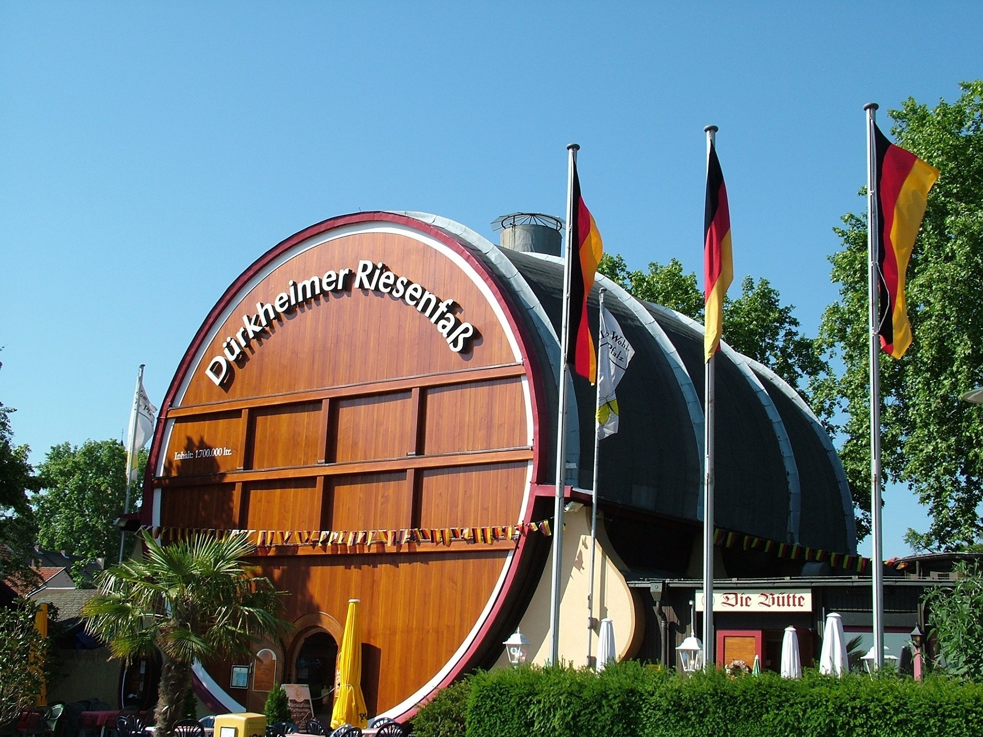 Das Dürkheimer Riesenfass Ansicht des von der Sonne angestrahlten dunkelbraunen Dürkheimer Riesenfasses in Bad Dürkheim vor strahlend blauem Himmel. Links unten im Bild sind eine Palme und rechts im Bild große grüne Bäume zu sehen, auf der rechten Seite wehen an Fahnenmasten drei Deutschland Fahnen.