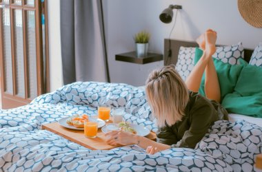 Eine junge Frau mit kurzen blonden Haaren, ihr Gesicht ist nach unten geneigt, liegt mit angewinkelten Beinen auf dem Bauch auf einem Bett mit blau weißer gemusterten Bettwäsche, vor ihr ein Tablett mit Frühstück und zwei mit  Orangensaft gefüllten Gläsern.