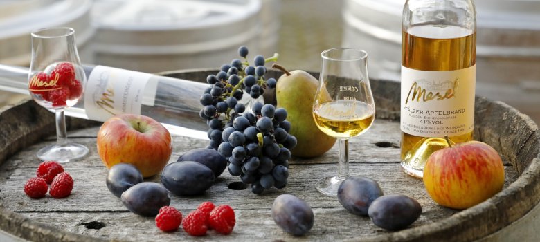 Brände mit Obst vom Weingut und Brennerei Mesel auf einem Weinfass präsentiert.