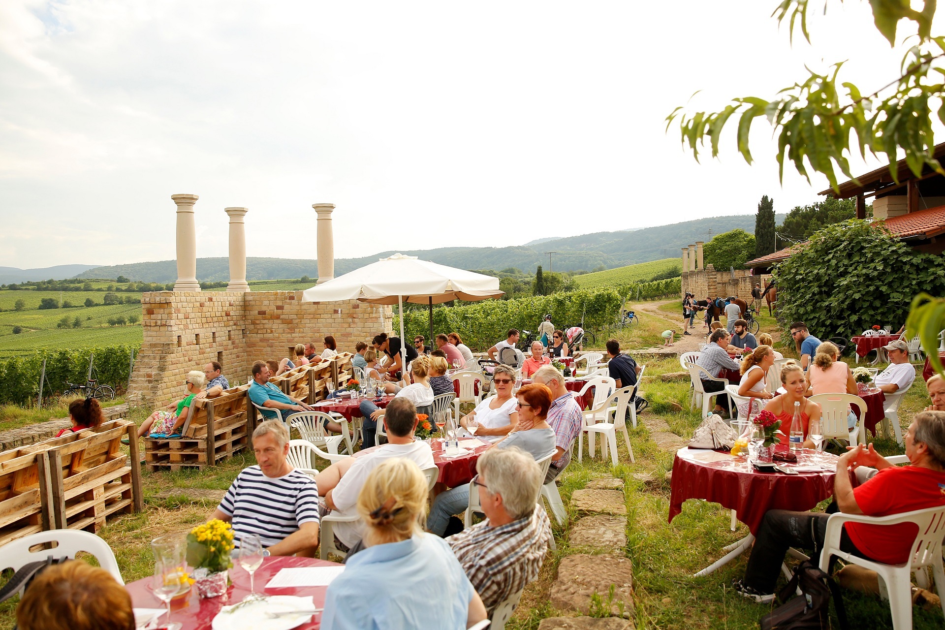 Veranstaltung unterhalb der Römervilla Weilberg Unterhalb der Römervilla Weilberg sitzen Personen auf Stühlen und auf Picknickdecken. Die haben Essen und Getränke vor sich. Im Hintergrund erkennt man die Säulen der Römervilla und die saftig grünen Weinberge.