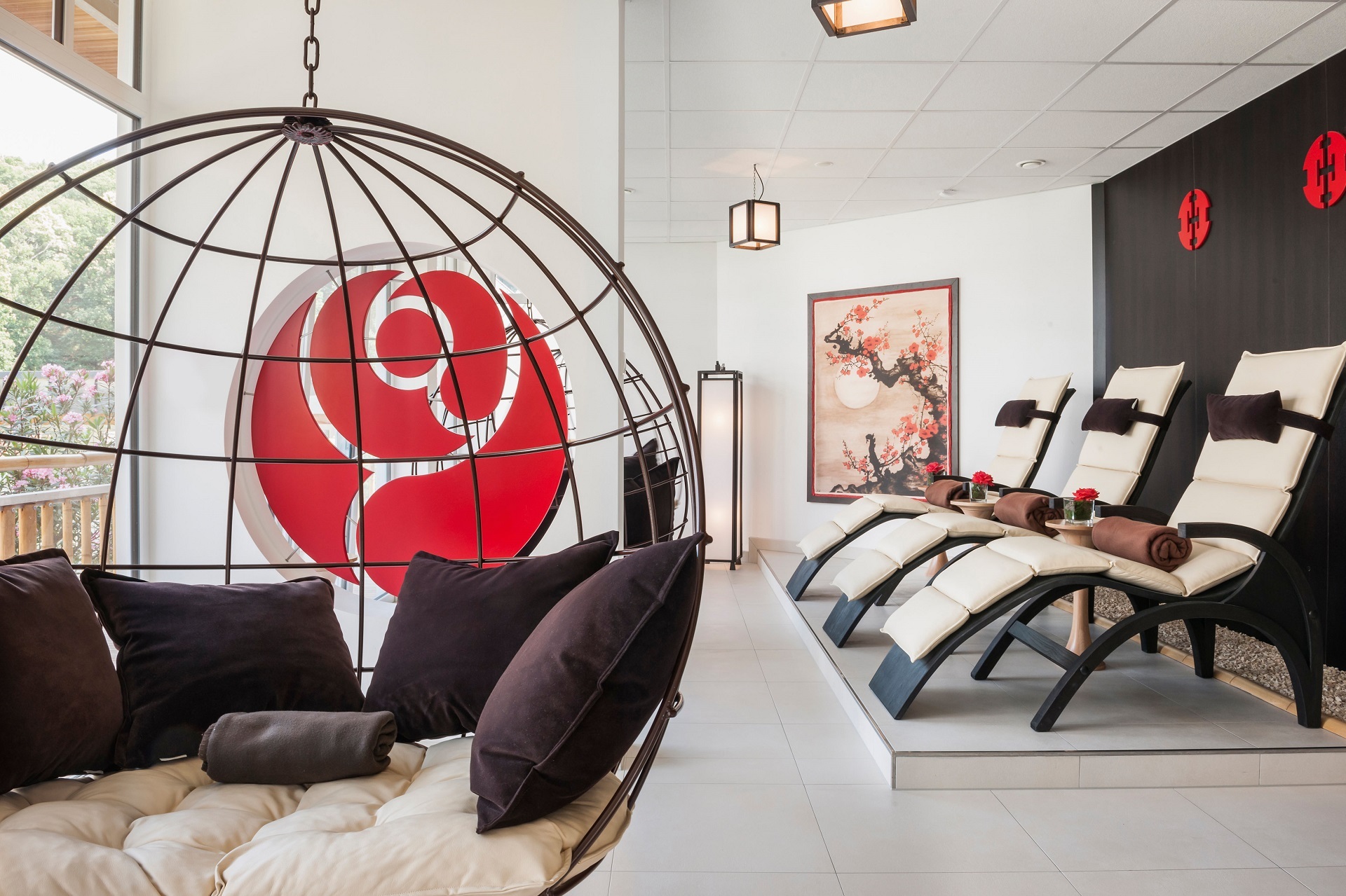 Zu sehen sind Liegestühle welche etwas erhöht im Ruhebereich vorzufinden sind. Vorne links befindet sich ein großer Runder Korb welcher von der Decke hängt. Er ist ausgestattet mit mehreren Kissen und einem gepolsterten Sitzbereich.