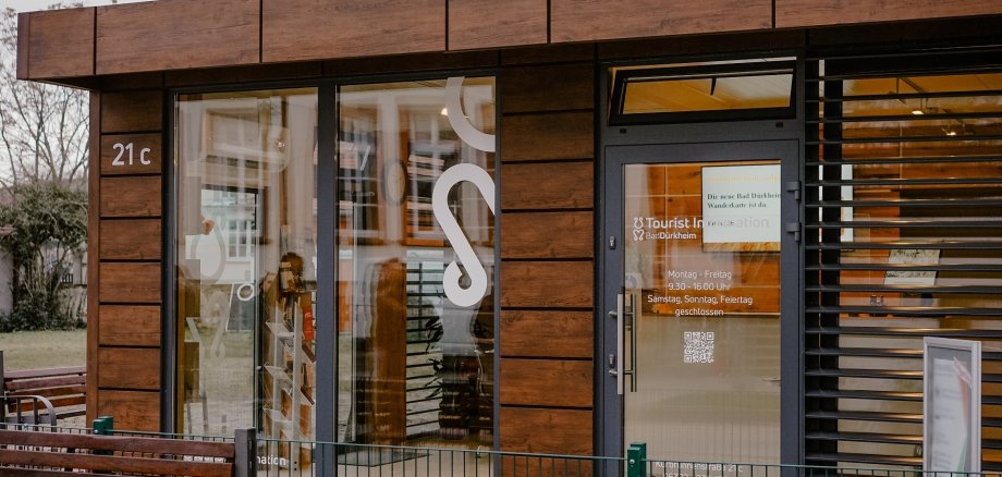 Eine Nahaufnahme der dunkelbraunen Außenfassade der Tourist Information mit zwei großen bodentiefen Fenstern und dem Eingangsbereich. Im Vordergrund ist ein niedriger Zaun und eine braune Holzsitzbank unscharf zu erkennen.