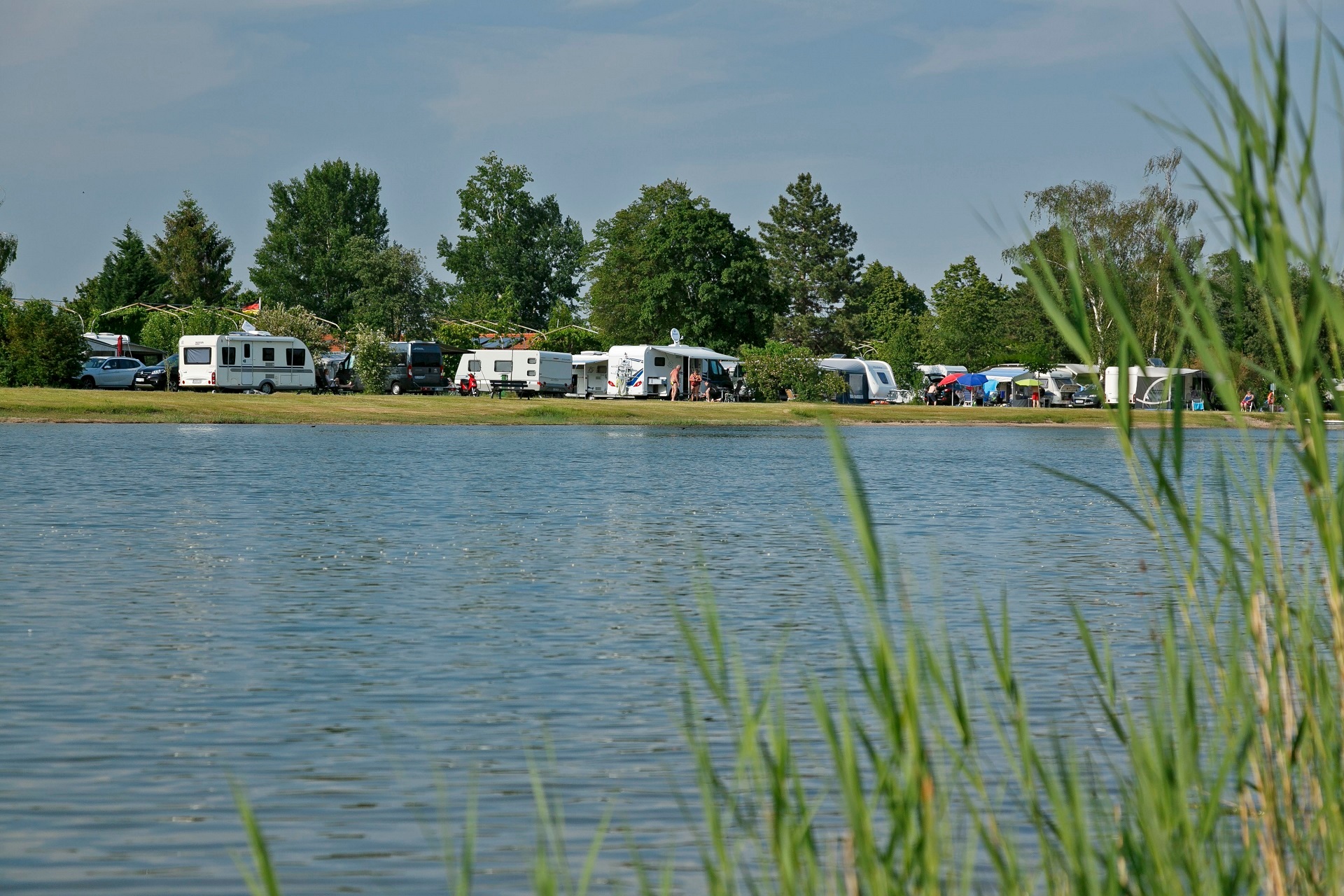 Im Vordergrund ist der Almensee und unscharf rechts im Vordergrund etwas Schild zu sehen. Im Hintergrund ist der Campingpark Knaus mit vielen parkenden Wohnmobilen zu sehen.