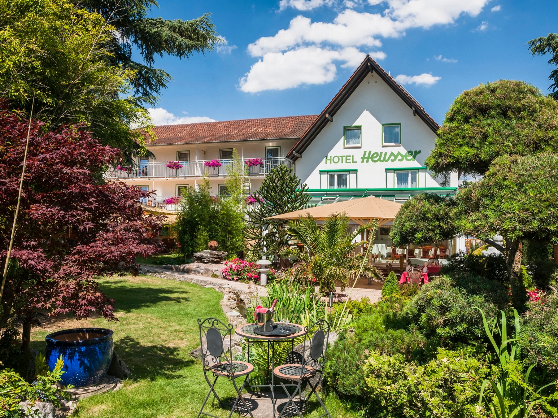 Ein Blick auf das Gartenhotel Heusser mit weißer Außenfassade und grüner Aufschrift "Hotel Heusser" , im Vordergrund der Garten des Hauses mit einer kleinen Sitzgruppe, gerahmt wird das Bild von Bäumen und Büschen.