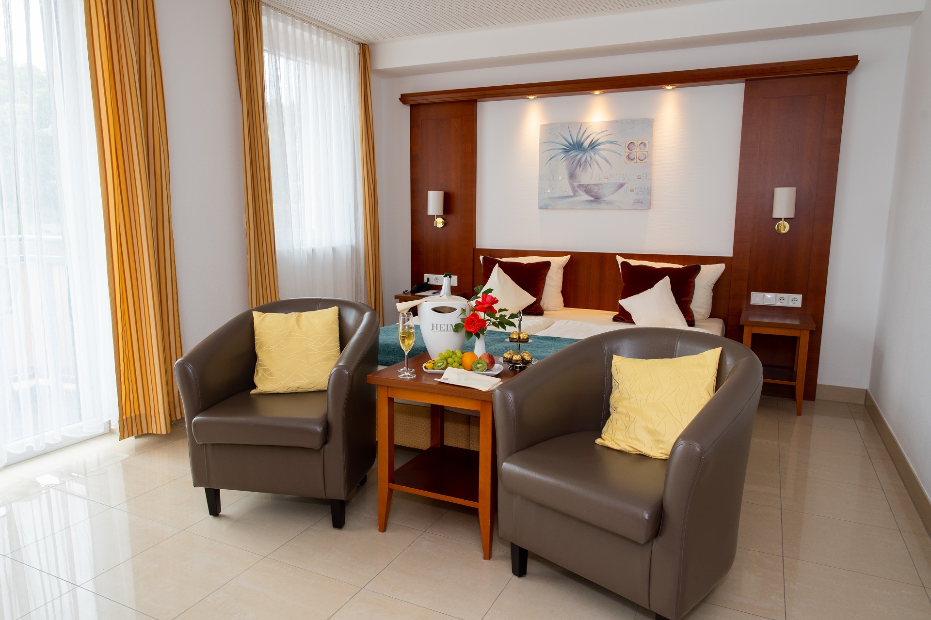 Ein Hotelzimmer im Gartenhotel Heusser mit weißen Bodenfließen, einer Holzfassade hinter dem Doppelbett, gelben Vorhängen und zwei braunen Sesseln am Fußende des Bettes mit einem Beistelltisch.