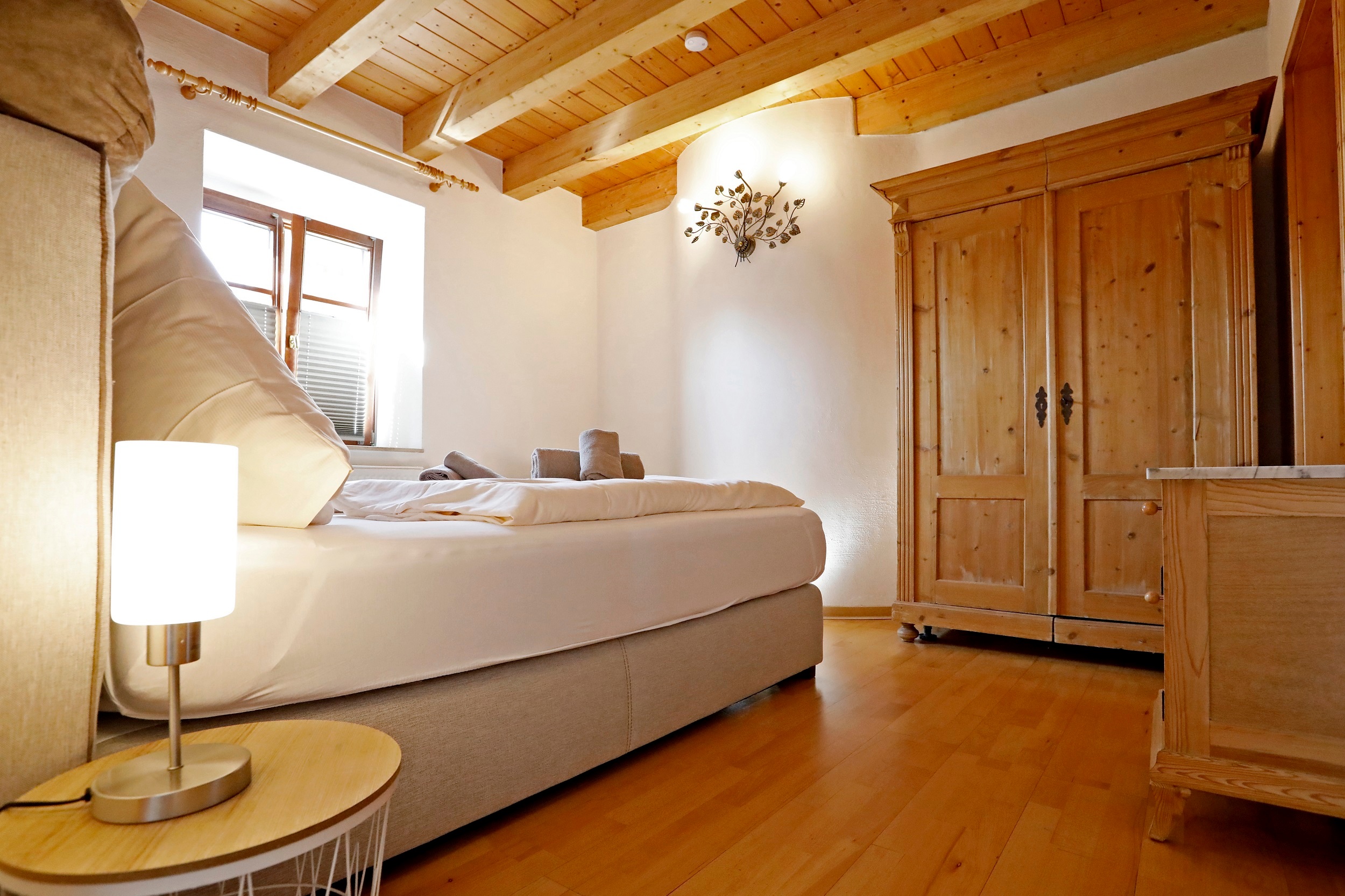 Ein Schlafzimmer der Ferienwohnung Gute Lage in Bad Dürkheim mit hellem Holzboden, in der rechten hinteren Ecke steht ein großer Holzkleiderschrank, links im Bild ist das beigene Doppelbett zu sehen. Die Wände strahlen in weiß und die Zimmerdecke ist ebenso aus hellem Holz mit Holzbalken.