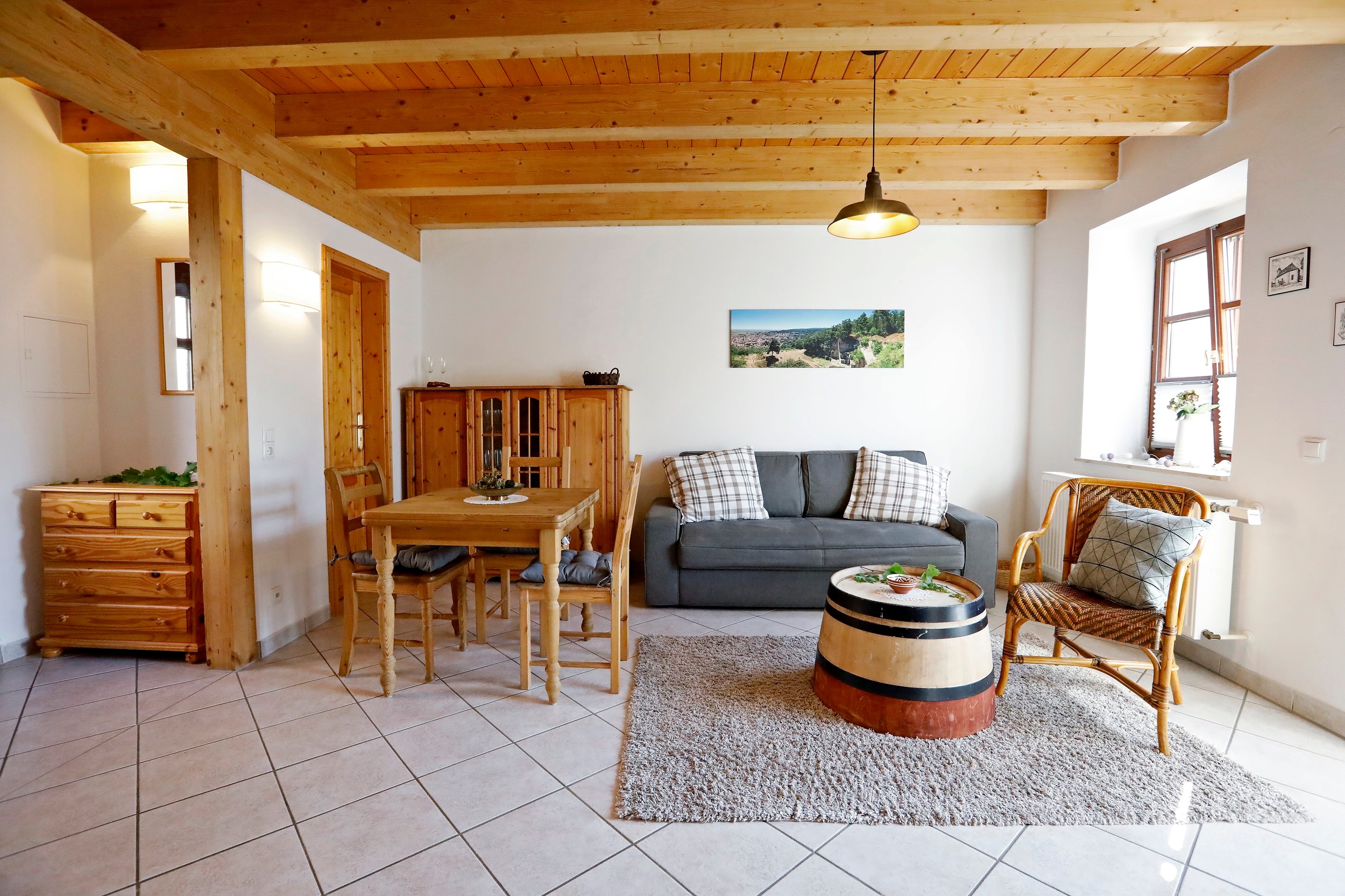 Der helle Wohnbereich der Ferienwohnung Gute Lage Nussriegel. Abgebildet ist hier rechts der Wohnbereich, ausgestattet mit einem blauen Zwei-Sitzer Sofa und einem braunen Sessel auf einem grauen flauschigen Teppich. Links im Bild ein kleiner Holztisch mit zwei Holzstühlen, im Hintergrund ein Holzschrank.