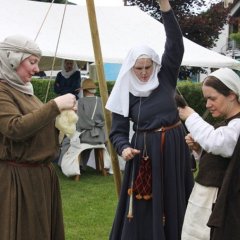 Zwei Frauen und ein Mann in mittelalterlichem Gewand stellen gemeinsam aus Wolle einen Garn her. Dazu zieht eine Frau die Wolle lang und die Andere verfilzt die Wolle bis eine Art Faden entsteht.