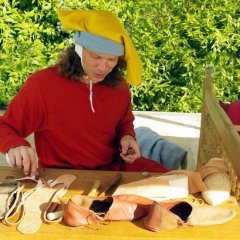 Zu sehen ist ein Schuhmacher mit knalligem roten Pullover und großer gelber Stoffmütze. Er hält Werkzeug in seiner linken Hand. Vor ihm liegen verschiedene Teile einen Stoff- oder Lederschuhs.
