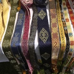 Eine Vielzahl an aufwendig verzierten Bändern aus Seide liegen auf einem weißen Tisch. Die Bänder sind an den Enden verknotet und geflochten. Sie haben unterschiedliche Farben und verschiedene Muster.