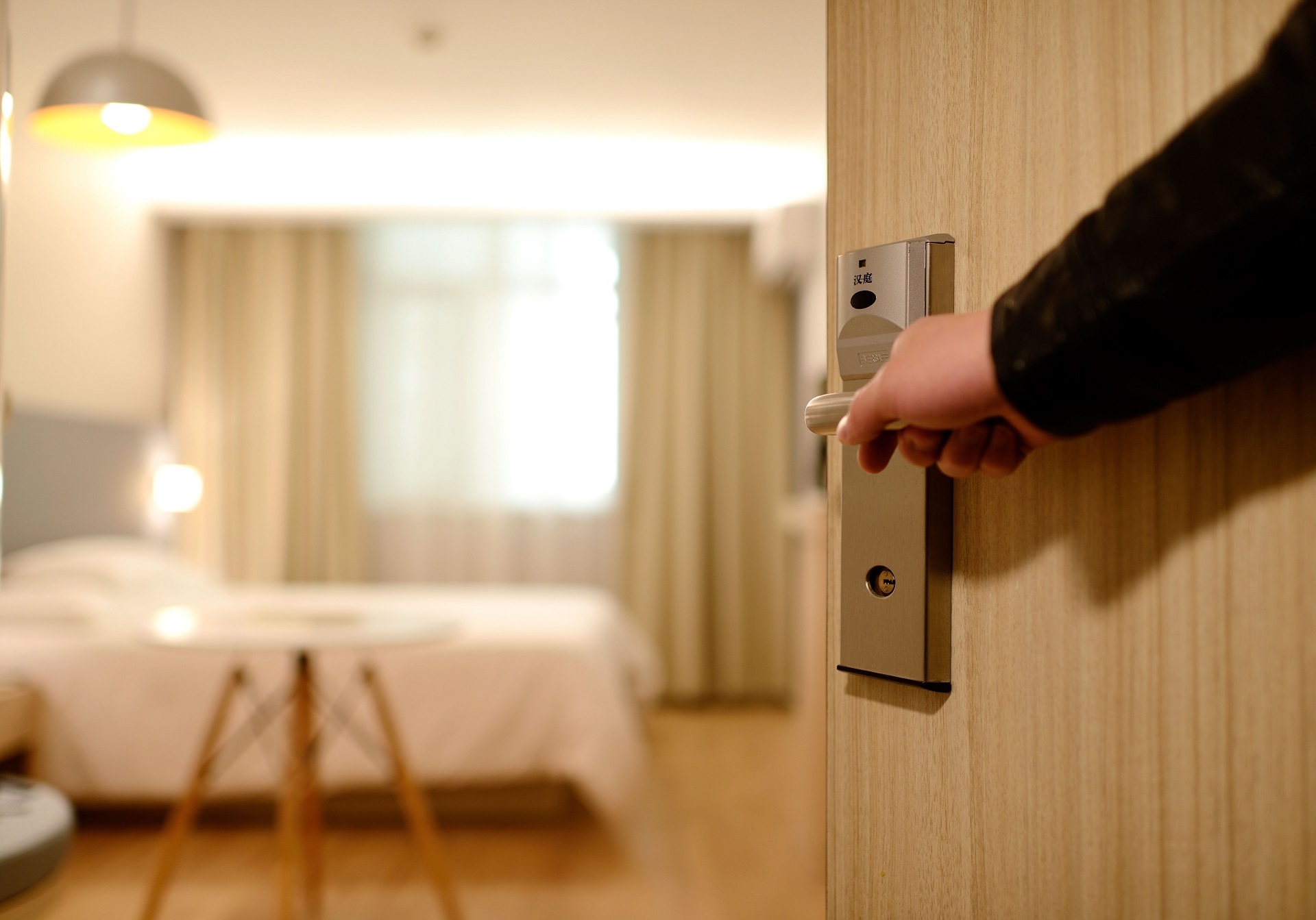 Im Vordergrund wird eine Hotelzimmer-Tür von einer Person mit schwarzen langen Ärmeln geöffnet, im Hintergrund ist ein Hotelzimmer mit einem mit weißer Bettwäsche bezogenem Bett, einer weißen Deckenlampe, sowie einem weißen Beistelltisch mit Holzfüßen zu sehen.