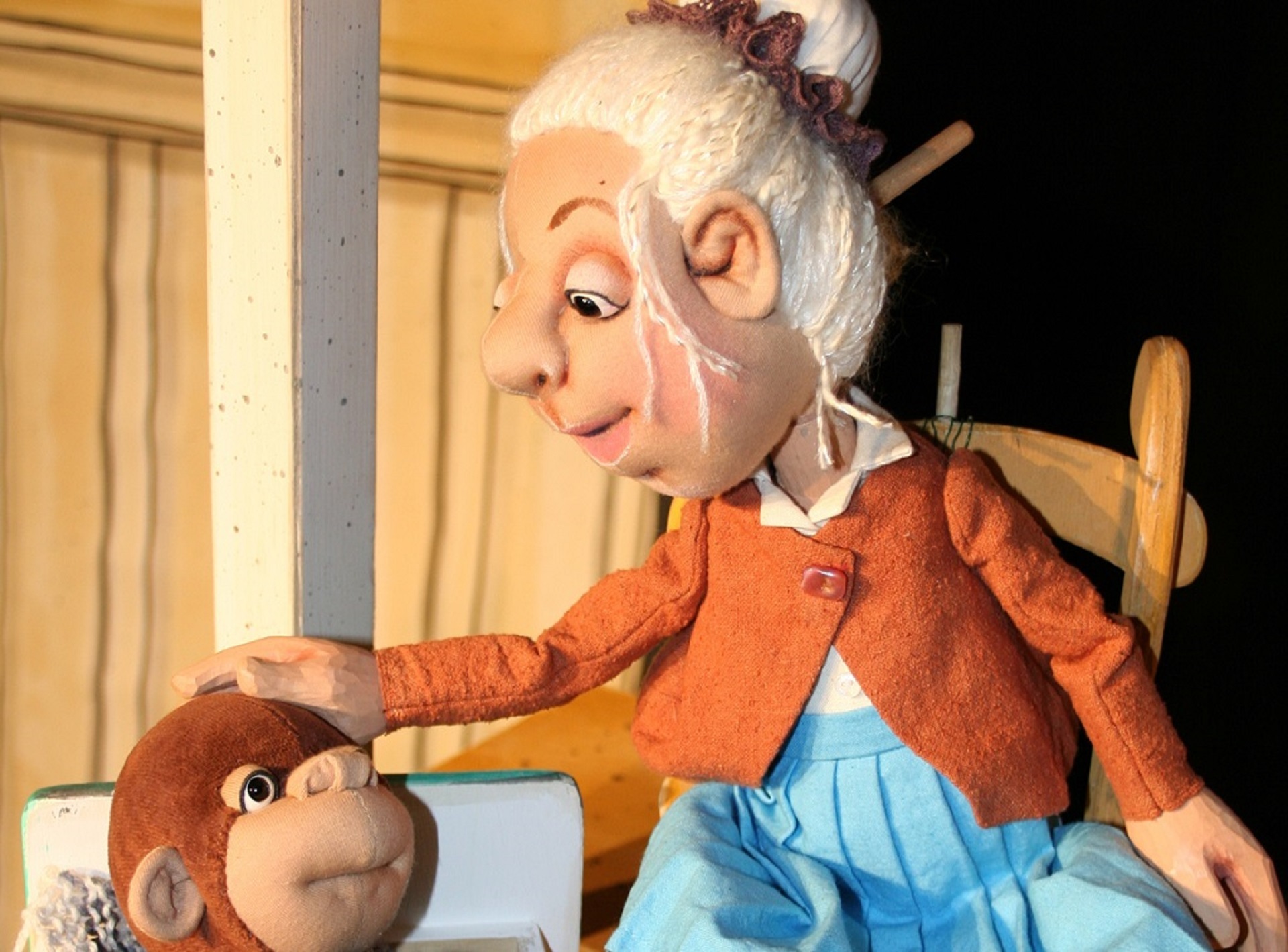 Im Fokus steht die Puppe "Oma Adele". Sie trägt weißes, in einem Dutt zusammengebundenes Haar, eine orangene Weste mit weißen Kragen und einen hellblauen Rock. Sie streichelt einen kleinen, brauen Stoff-Affen. Oma Adele sitzt auf einem brauen Holzstuhl, während der Affe aus einen kleinen Holzkiste ragt.