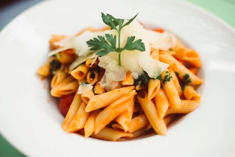 In einem vertieften Teller ist ein Pastagericht liebevoll angerichtet. Das Gericht besteht auf Penne Nudeln in Tomatensoße mit Spinat und Parmesan. Angerichtet ist dies mit Petersilie.