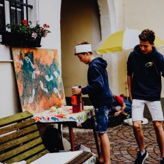 Zwei Jungs bauen rote Dosen im Innenhof des Haus Catoirs auf, um Dosenwerfen spielen zu können. Der Innenhof ist durch verschiedene, selbst gemalte Bilder geschmückt.