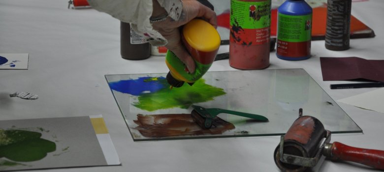 Verschiedene Kunstwerke auf Glasplatten gemalt liegen auf einem Tisch. Dazwischen stehen Acrylfarben, Farbrollen und Farbpaletten.