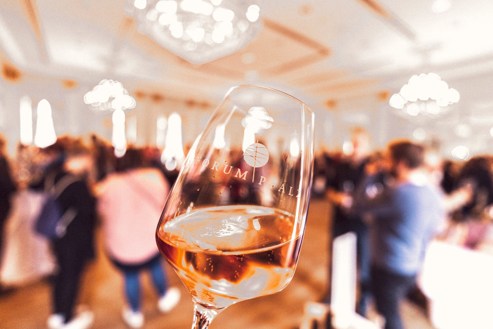 Im Vordergrund ist ein Stielglas mit dem Logo des Forum Pfalz zu sehen, im Hintergrund die Veranstaltung Forum Pfalz selbst, mit einigen Besuchern im Kurhaus