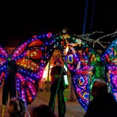 Zwei als leuchtende Schmetterlinge verkleidete Stelzendarstellerinnen flankieren ihren mittig laufenden Kollegen, der einen Zylinder, Anzug und einen Stab trägt, dessen Spitze leuchtet.