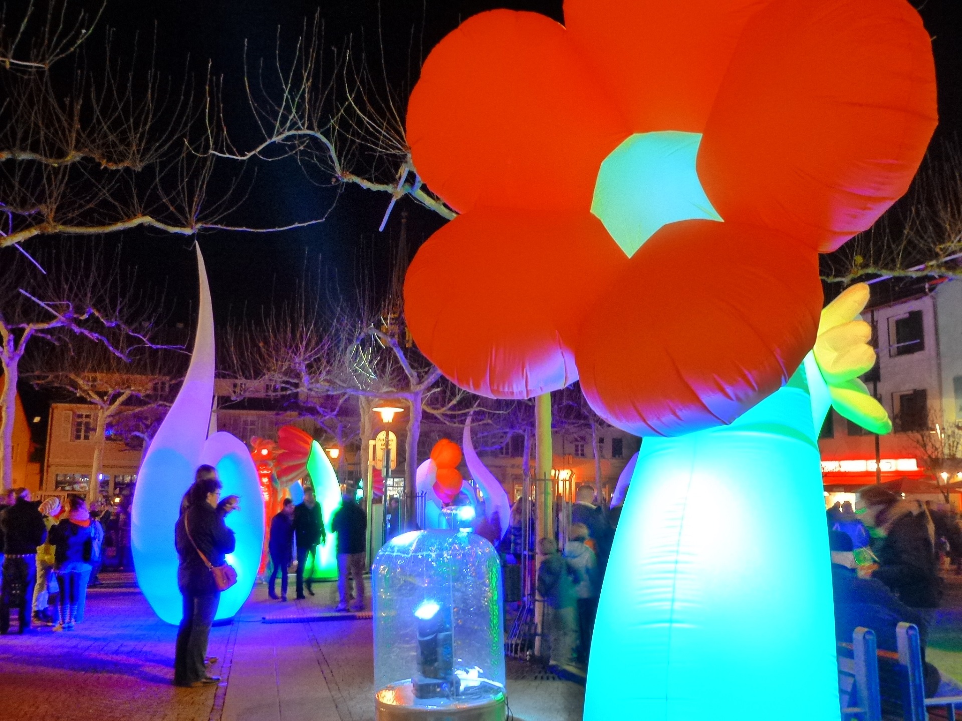 Im Vordergrund eine Leuchtfigur in Form einer roten Blume, im Hintergrund Besucher der Bad Dürkheimer (W)Einkaufsnacht und weitere bunt beleuchtete Leuchtkegel die den Stadtplatz schmücken