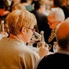 Zu sehen ist aus der Perspektive hinter einer Dame, wie sie an einem Stielglas mit Rotwein gefüllt riecht, um sie herum sitzen viele Besucher der Bad Dürkheimer Weinkür