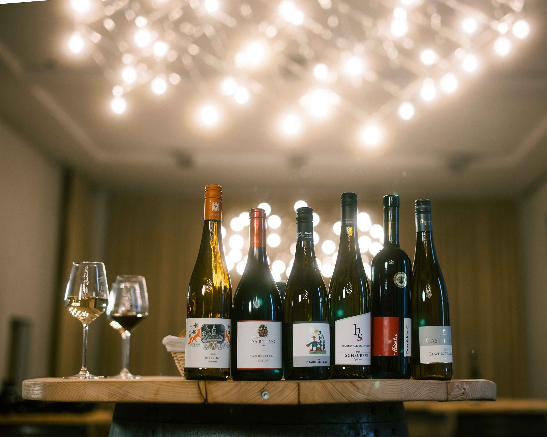 Eine Frontalaufnahme von sechs unterschiedlichen Weinflaschen nebeneinander gereiht und zwei Stielgläser daneben. Im Hintergrund ist die Decke der Brunnenhalle zu sehen und eine funkelnde Leuchte.