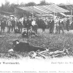 Bei dem Bild handelt es sich um eine alte Postkarte vom Wurstmarkt im Jahr 1902. Zu sehen sind ein Teil der damaligen Brühlwiesen und die damaligen Schubkarchstände, die von Planen geschützt werden. Im Vordergrund liegen zwei Männer auf dem Boden die als "Zwei Opfer des Wurstmarktes" bezeichnet werden. Hinter ihnen steht eine kleinere Ansammlung an Menschen, die sie beobachtet.