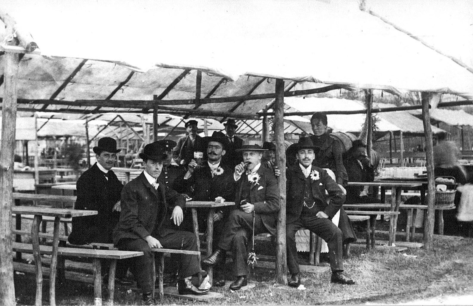 Im Vordergrund des Bildes sind sechs Männer zu sehen, die in Anzügen und schicken Hüten wie Melonen gekleidet sind. Sie sitzen in einem der vielen Schubkarchstände auf dem Wurstmarkt von 1913 und genießen Zigarren und Wein. Im Hintergrund ist eine Frau und einzelne Männer zu sehen. Es ist zu erkennen, dass die damaligen Versionen der Schubkarchstände noch viel schmaler als heutzutage sind.