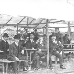 Im Vordergrund des Bildes sind sechs Männer zu sehen, die in Anzügen und schicken Hüten wie Melonen gekleidet sind. Sie sitzen in einem der vielen Schubkarchstände auf dem Wurstmarkt von 1913 und genießen Zigarren und Wein. Im Hintergrund ist eine Frau und einzelne Männer zu sehen. Es ist zu erkennen, dass die damaligen Versionen der Schubkarchstände noch viel schmaler als heutzutage sind.