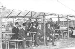 Im Vordergrund des Bildes sind sechs Männer zu sehen, die in Anzügen und schicken Hüten wie Melonen gekleidet sind. Sie sitzen in einem der vielen Schubkarchstände auf dem Wurstmarkt von 1913 und genießen Zigarren und Wein. Im Hintergrund ist eine Frau und einzelne Männer zu sehen. Es ist zu erkennen, dass die damaligen Versionen der Schubkarchstände noch viel schmaler als heutzutage sind.