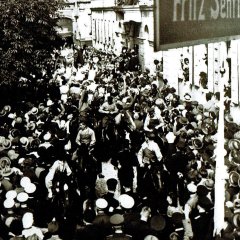 Zu sehen ist eine große Menschenmasse, die am Wurstmarktumzug von 1924 teilnimmt. Die Masse stellt sich auf dem Schlossplatz auf. Es sind ebenfalls Personen zu sehen, die den Umzug aus ihren Fenstern heraus beobachten.