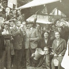 Zu sehen ist eine größere Gruppe von Menschen an einem Schubkarchstand im Jahr 1931. Einer der Männer vorne im Bild sitzt auf dem Boden und hält einen Eimer im rechten Arm und deutet mit der linken Hand zur Kamera. Einige der Leute machen lustige Gesichter und stoßen mit Dubbegläsern an. 