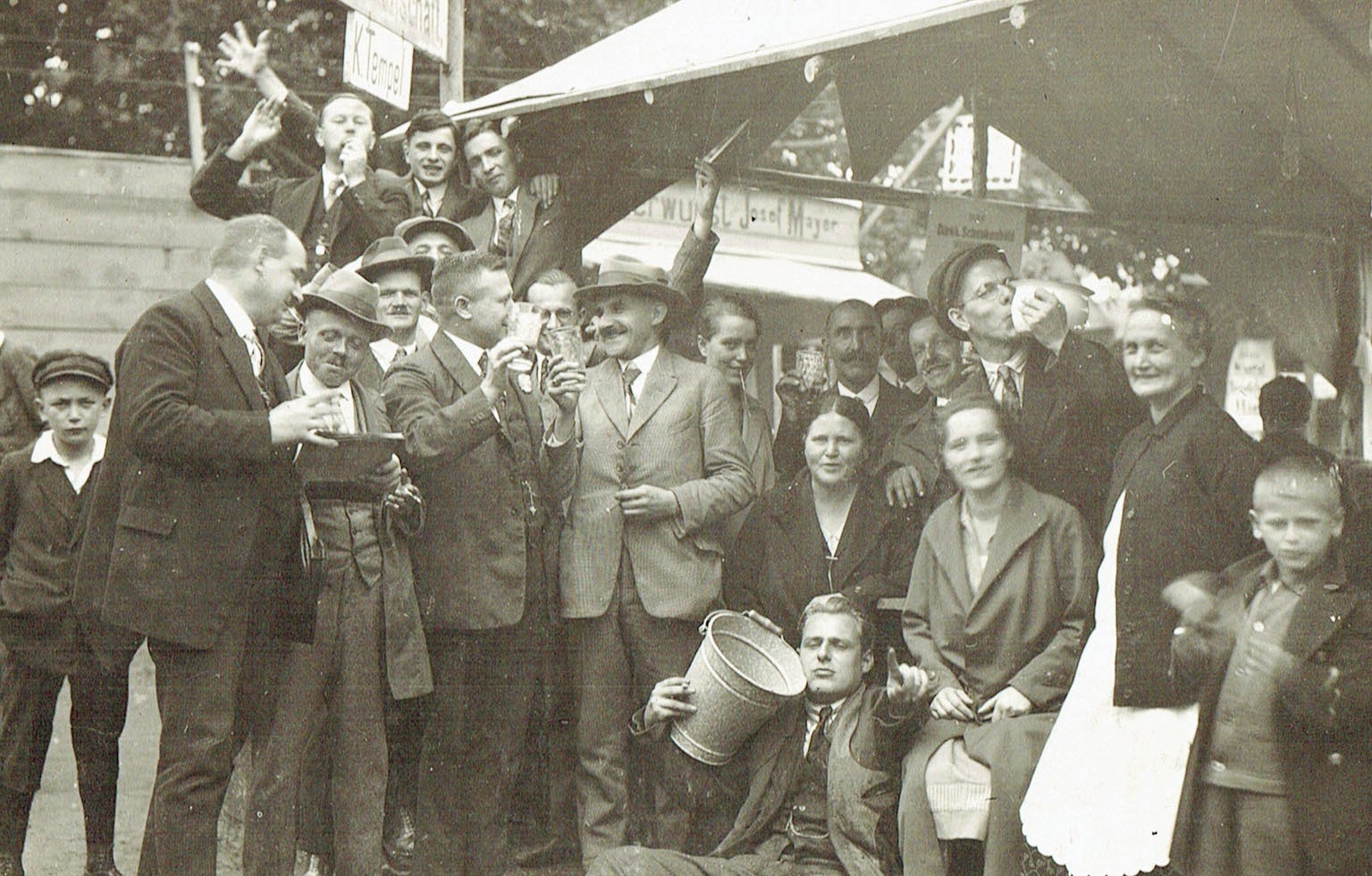 Zu sehen ist eine größere Gruppe von Menschen an einem Schubkarchstand im Jahr 1931. Einer der Männer vorne im Bild sitzt auf dem Boden und hält einen Eimer im rechten Arm und deutet mit der linken Hand zur Kamera. Einige der Leute machen lustige Gesichter und stoßen mit Dubbegläsern an. 