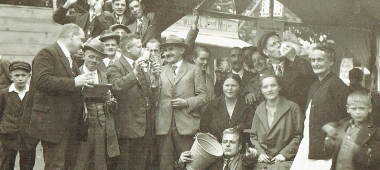 Zu sehen ist eine größere Gruppe von Menschen an einem Schubkarchstand im Jahr 1931. Einer der Männer vorne im Bild sitzt auf dem Boden und hält einen Eimer im rechten Arm und deutet mit der linken Hand zur Kamera. Einige der Leute machen lustige Gesichter und stoßen mit Dubbegläsern an. 