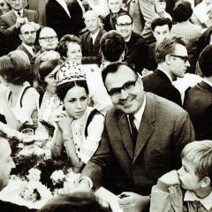 Nach der Wurstmarkteröffnung sitzt eine Gruppe von Menschen an einem Tisch. Im Fokus des Bildes steht Helmut Kohl, der einen Anzug und Brille trägt, und direkt in die Kamera schaut und lächelt. Rechts von ihm aus sitzt eine ehemalige Weinkönigin, die ihre traditionelle Tracht und Krone trägt.