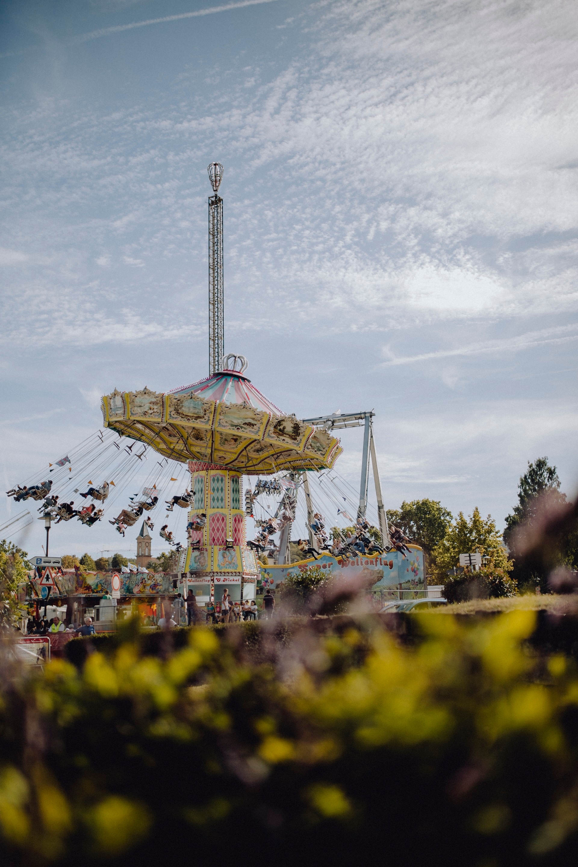 Bei schönem Wetter wird das alte Kettenkarussell auf dem Wurstmarktplatz von der Ferne abgebildet. Es befindet sich mitten in der Fahrt und Leute sind auf den einzelnen Sitzplätzen zu erkennen.