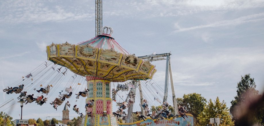 Bei schönem Wetter wird das alte Kettenkarussell auf dem Wurstmarktplatz von der Ferne abgebildet. Es befindet sich mitten in der Fahrt und Leute sind auf den einzelnen Sitzplätzen zu erkennen.
