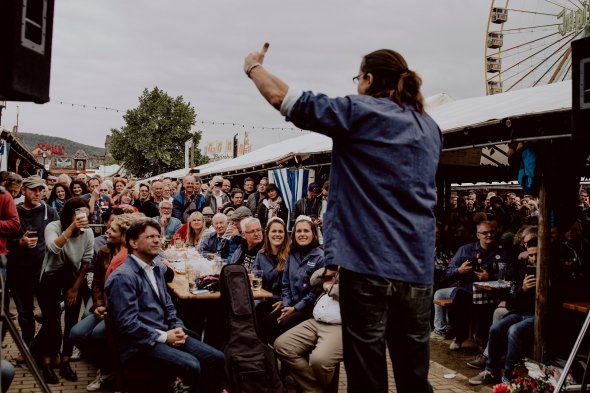 Comedian Chako Habekoste steht mit dem Rücken zur Kamera gewandt auf der kleinen Bühne des literarischen Frühschoppens auf dem Dürkheimer Wurstmarkt und unterhält die Menschen die ihm gegenüber stehen und sitzen.