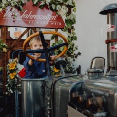 Ein kleiner Junge mit Winzerkittel bekleidet, sitzt auf dem Traktor welcher den Vier Jahreszeiten Wagen zieht. Der Junge schaut zwischen dem großen Lenkrad hervor.
