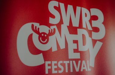 Ein weißer Schriftzug "SWR3 Comedy Festival" befindet sich auf rotem Grund. Es wurde seitlich fotografiert.