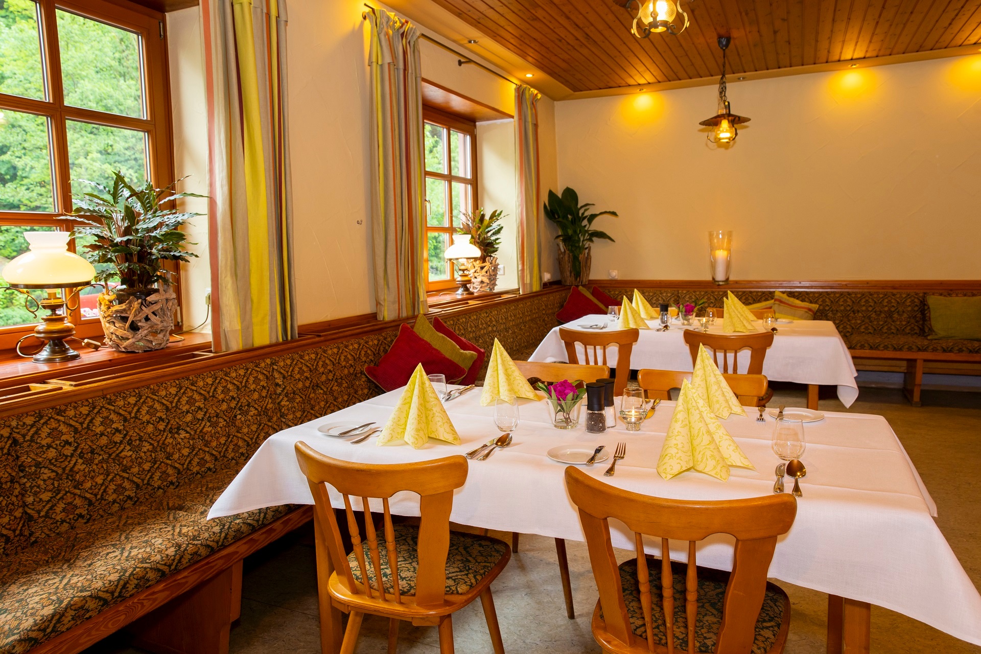 Innenbereich der Waldgaststätte Alte Schmelz. Zu sehen sind zwei hintereinander stehende eingedeckte Tische. An den Tischen verläuft unter den zwei Fenstern auf der linken Seite eine lange gemütliche Eckbank.